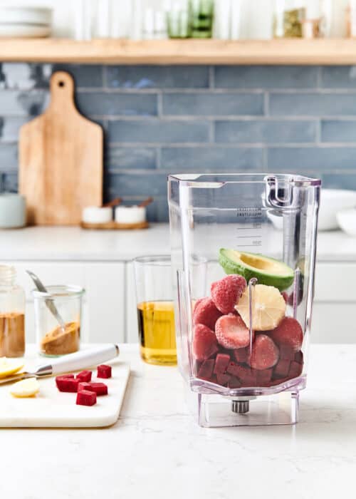 Blender full of beets, strawberries & avocado with ingredients on a wood cutting board in a white marble kitchen