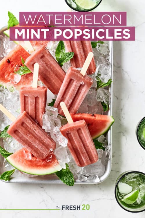 Sheetpan full of fresh watermelon and homemade watermelon mint popsicles with ice on a white marble surface