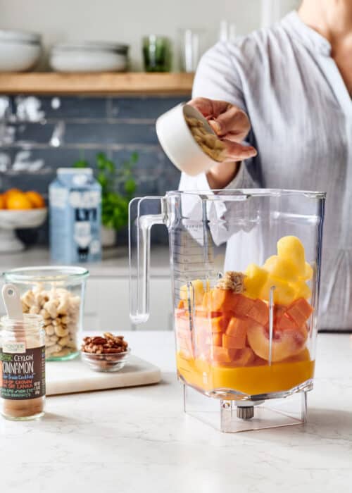 Woman pouring nuts into a blender full of carrots, peaches, pineapple & orange juice in a white marble kitchen