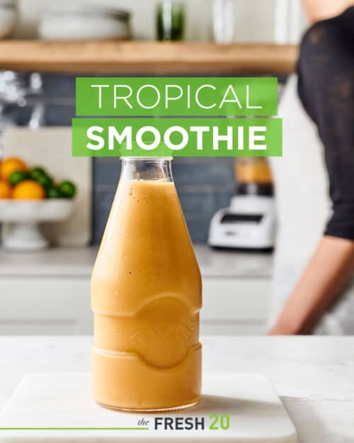 Woman making a full container of fresh mango, banana, & papaya tropical smoothie in a beautiful white marble kitchen