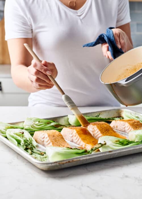 Woman glazing a sheet pan full of maple soy salmon and bok choy on a white marble surface