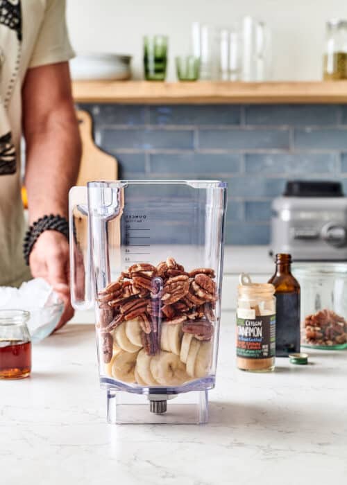 Man making a blender full of bananas, pecans, cinnamon, honey & vanilla in a white marble kitchen