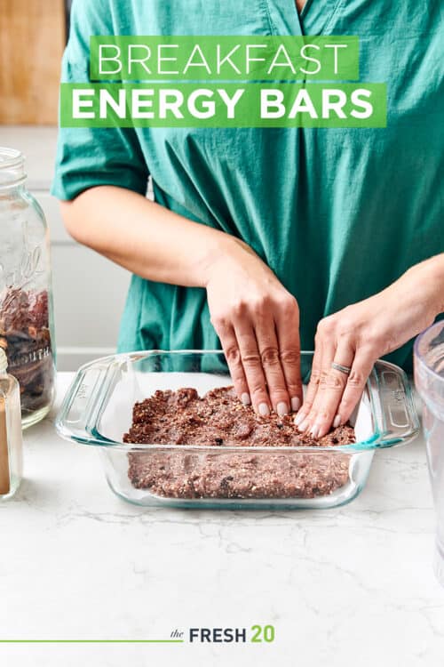 Woman pressing her hands into no bake breakfast bar batter into a glass baking dish next to jars of ingredients in a beautiful white marble kitchen