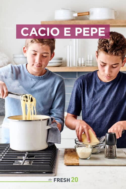 Two boys cooking pasta noodles in a white stockpot & grating parmesan cheese in a white marble kitchen