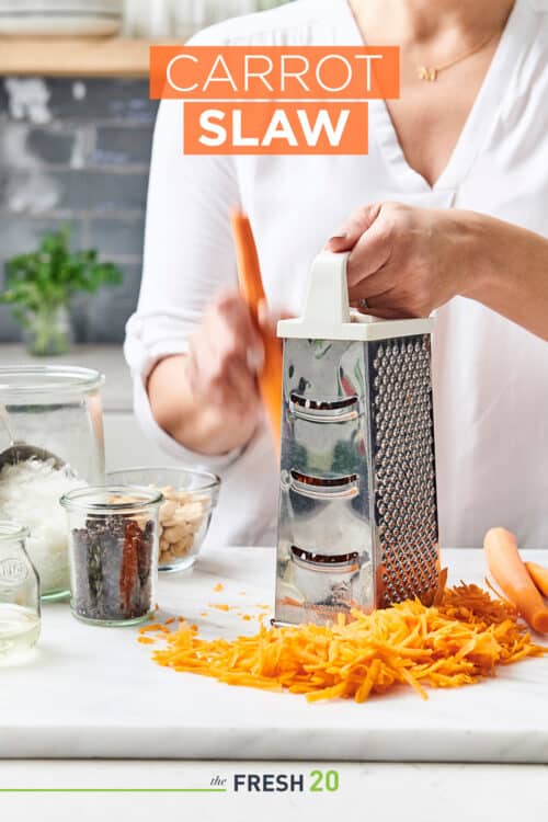 Woman shredding carrots with a metal box grater onto a white marble surface with raisins, coconut & nuts