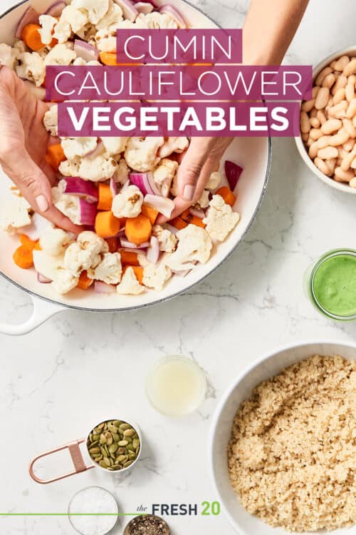 Hands tossing an easy roasted cauliflower cumin vegetable salad in a white skillet with bowls of ingredients on a white marble counter