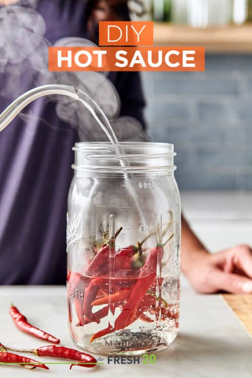 Woman pouring steaming boiling water from a kettle into a mason jar filled with deep red hot chili peppers