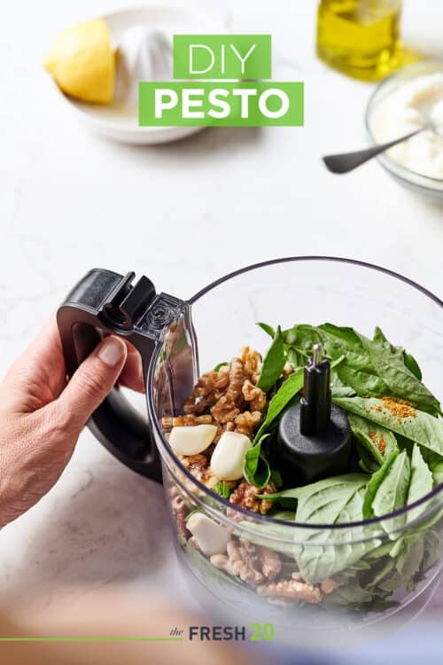 Food processor full of fresh basil, garlic and walnuts along with a lemon juicer & olive oil on a white marble surface