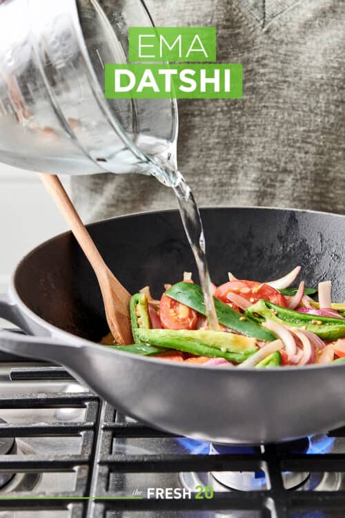 Pouring water into a Le Creuset wok filled with chilis, onions & tomatoes on a gas cooktop