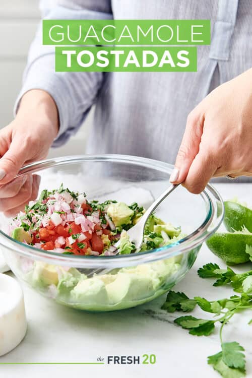 Woman mashing avocados, tomatoes & onions with a fork in a glass bowl for guacamole with lime and cilantro on a white marble surface