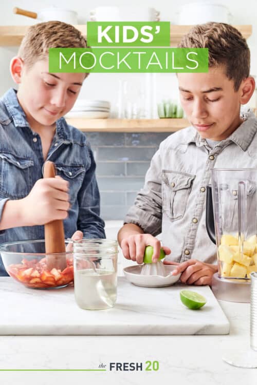 2 young boys muddling strawberries & juicing limes with a pitcher of pineapple in a white marble kitchen