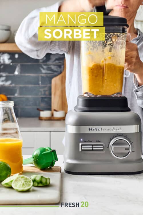 Woman using KitchenAid blender filled with mangos, limes and orange juice in a white marble kitchen