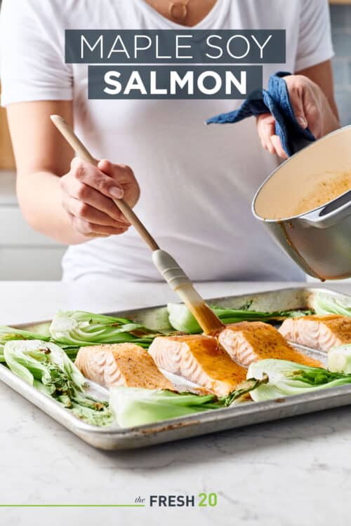 Woman glazing a sheet pan full of maple soy salmon and bok choy on a white marble surface