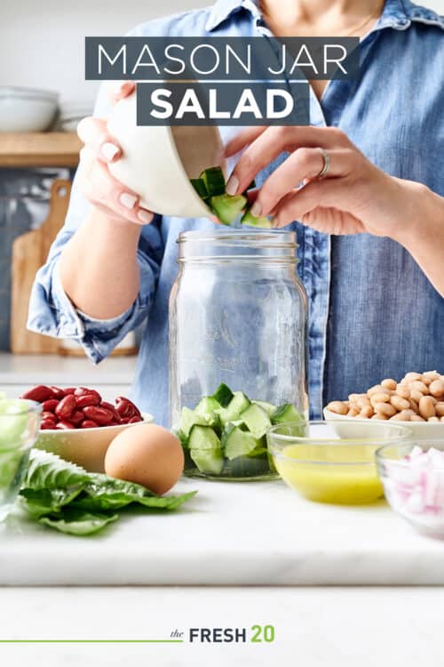 Woman filling glass mason jar with salad & bowls of vegetable ingredients in a white marble kitchen