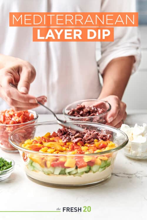 Woman using a spoon to layer olives into a serving bowl to create an easy 7-layer Mediterranean dip for entertaining