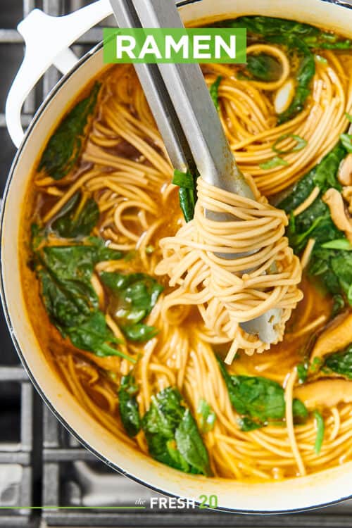 Silver tongs grabbing hearty ramen noodles from stockpot full of noodles, spinach and onions on a metal cooktop