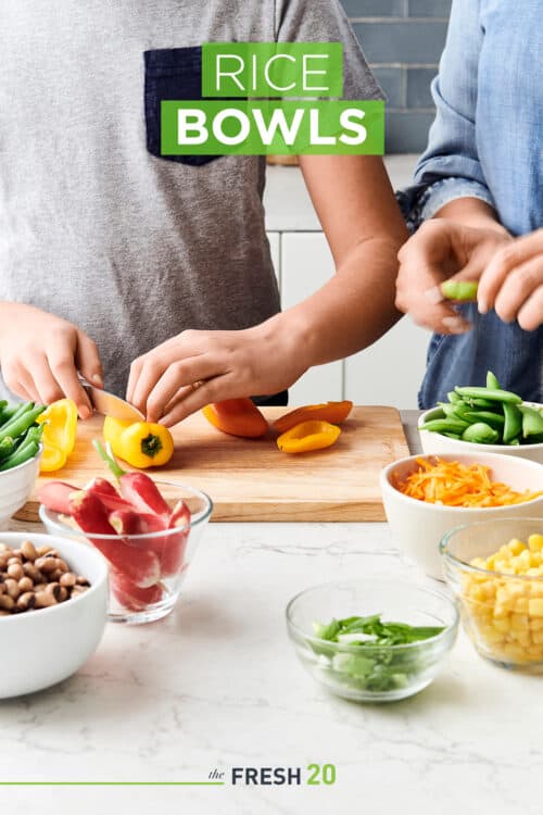 Two kids cutting fresh vegetables on a wooden cutting board with ingredients in bowls in a beautiful white marble kitchen