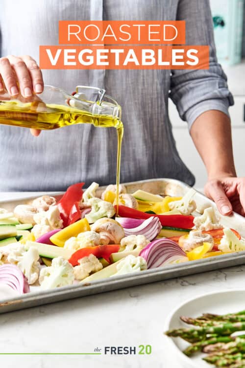 Woman pouring olive oil on a baking sheet filled with an assortment of vegetables on a white marble countertop