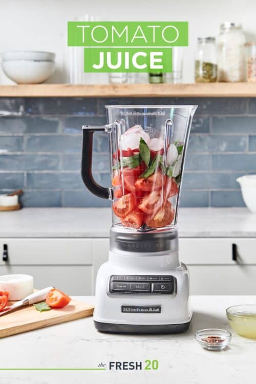 Blender full of tomatoes, basil & red bell peppers next to chopped ingredients on a wood cutting board in a white marble kitchen