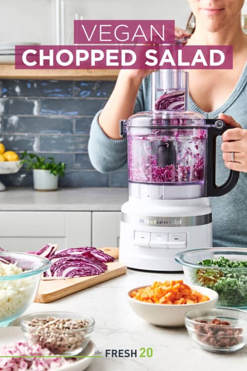Woman chopping red cabbage in food procesor surrounded with bowls of fresh cut vegetables for a vegan chopped salad in a beautiful white marble kitchen