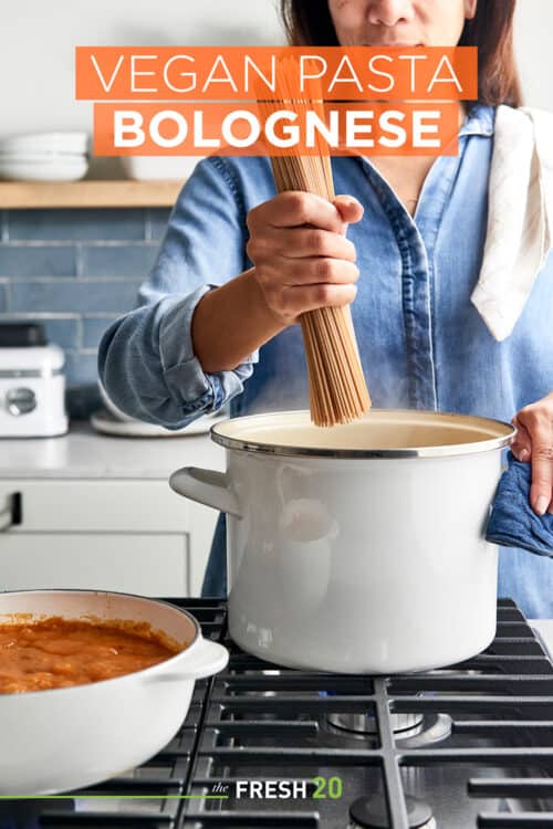 Woman placing uncooked pasta noodles into a cream colored pot next to pasta sauce on a metal stovetop