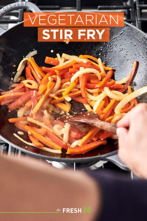 Le Creuset wok filled with carrots, onion and bell peppers being stirred with a wooden spatula on a cooktop