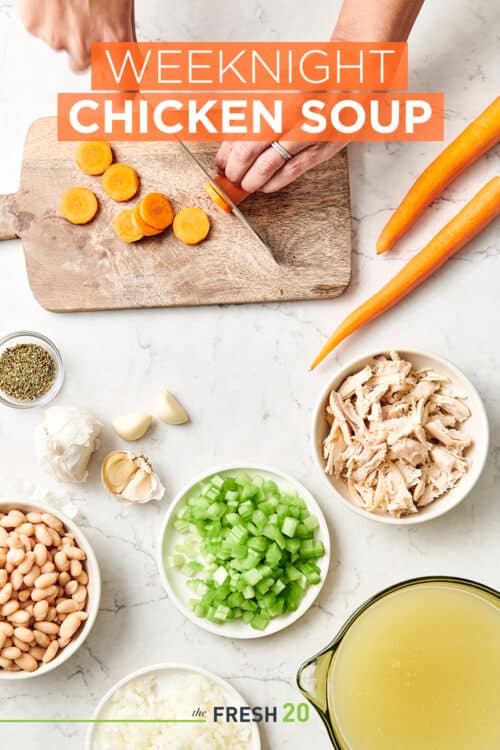 Woman cutting carrots on a wood cutting board with ingredients to make chicken soup on a white marble countertop