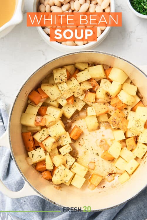 Le Cruset pot full of cubed potatoes and carrots alongside a bowl of white beans on a white marble surface