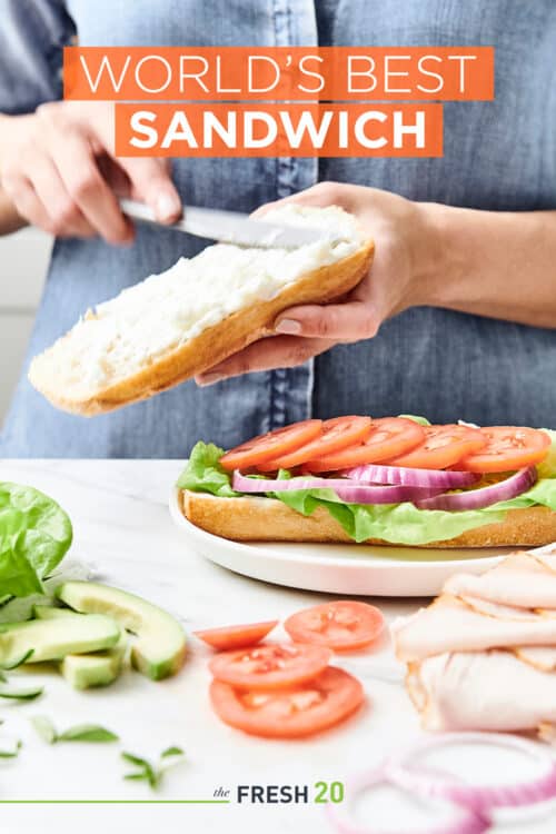 Woman spreading mayo on a layered turkey sandwich next to a variety of fresh ingredients on a white marble surface