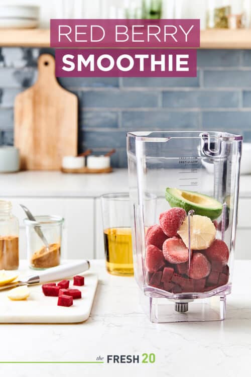 Blender full of beets, strawberries & avocado with ingredients on a wood cutting board in a white marble kitchen