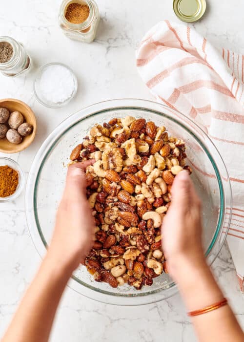 Hands mixing spiced nuts in a glass bowl on a white marble surface for the holidays