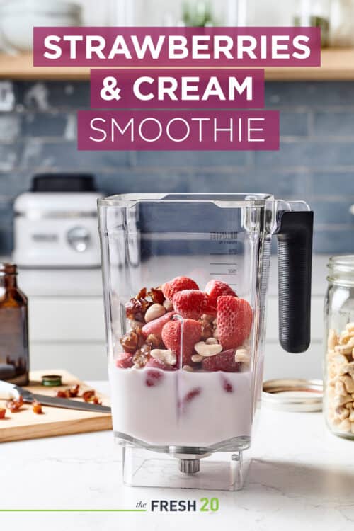 Blender full of strawberries, almond milk, nuts, dates & other ingredients on a wood cutting board in a white marble kitchen