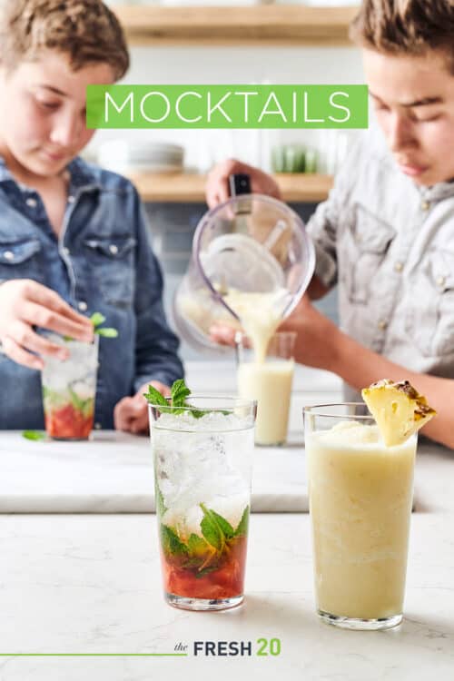 2 boys making 2 fresh fruit summer mocktails with mint leaves & pineapple garnish in a white marble kitchen