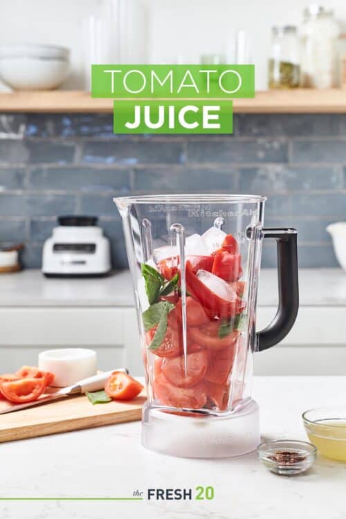 Blender full of tomatoes, basil & red bell peppers next to chopped ingredients on a wood cutting board in a white marble kitchen